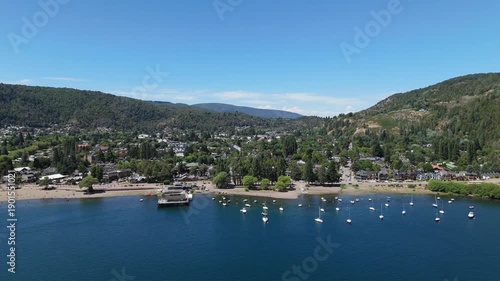 Aerial view of Lake Lacar and San Martin de los Andes in Patagonia, highlighting a scenic marina with numerous sailboats and boats anchored in deep blue water surrounded by mountains.