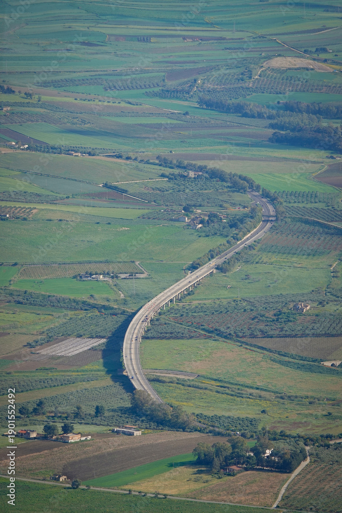 Fototapeta premium Long highway viaduct bending through green agricultural fields in western Sicily. The sweeping curve highlights modern infrastructure cutting across rural orchards and farmland on Mediterranean island