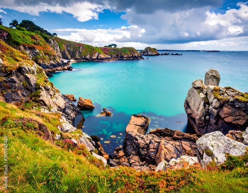 A vibrant coastal scene presents turquoise waters cradled by rocky cliffs covered in lush vegetation under a cloudy blue sky
