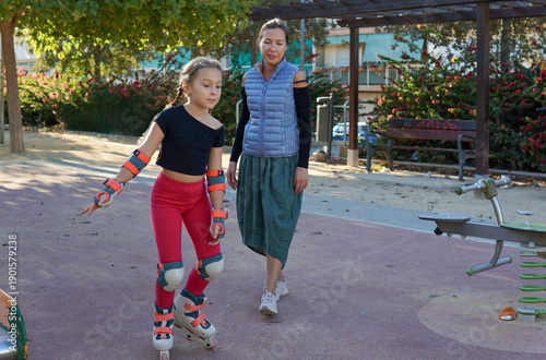 Mother teaching her young daughter to roller skate at a playground, holding her for support, having fun together outdoors.