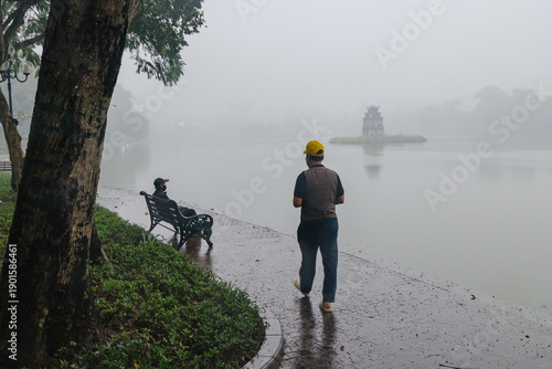 Two men on a foggy morning at Hoan Kiem Lake, Hanoi, Vietnam. One walks, another sits by the misty lake, with the iconic Turtle Tower in the distance.