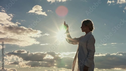 Silhouette of a woman in a light jacket holding her hand out towards the bright sun in a cloudy sky. Concept of hope, freedom, and connection with nature at sunset.