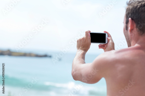 Adult male standing shirtless on shore holding phone framing ocean and rocky headland, copy space