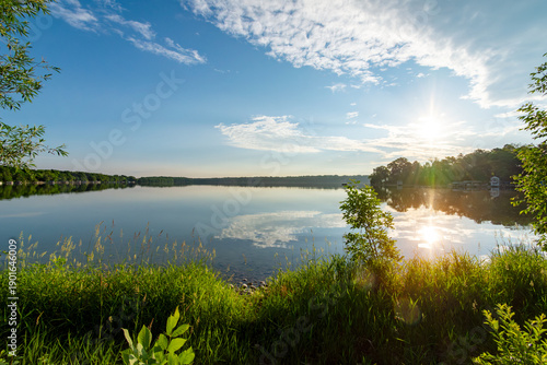 Lush green shoreline of a Waukesha County lake in early June. Bright morning sun reflects on calm Wisconsin water under blue skies.