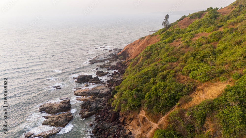 Obraz premium Aerial view of Arambol Beach, Goa, showing Arambol Mountain on the right with rugged rocks below as winter evening waves roll in from the left along the Arabian Sea coast.