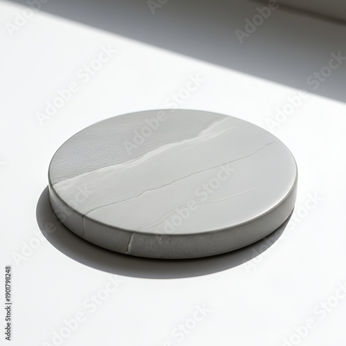 A round marble plate casts a shadow on a white surface, illuminated by natural light from an unseen source, creating a subtle gradient of light and shadow.