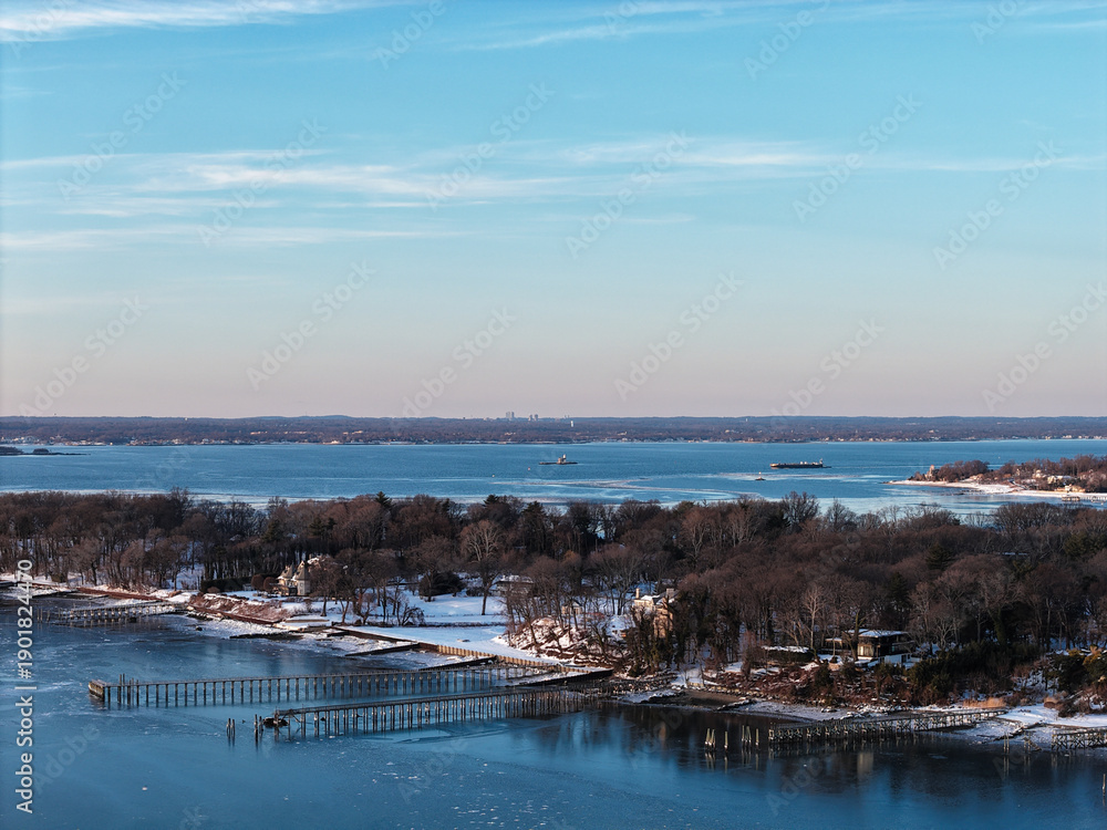 Obraz premium Long island sound shoreline with snow covered trees and frozen water near port washington, new york featuring a distant cityscape horizon on a clear winter day