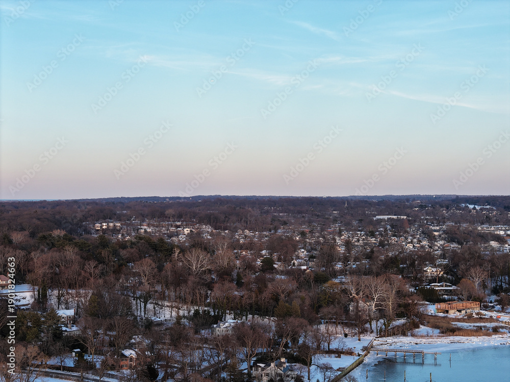 Obraz premium Aerial view displaying a peaceful winter landscape over port washington, new york, with suburban homes nestled among bare trees and snow lining the waterfront along plum beach point drive