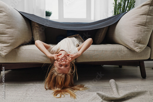 Playful child lies upside down under a blanket draped on a beige sofa in a bright living room. The kid smiles and covers eyes with hands creating a cozy and joyful indoor moment.