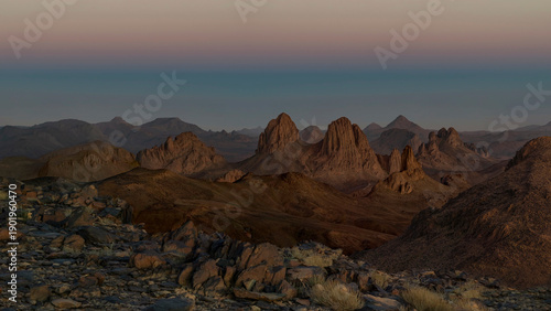 Elevated view from Assekrem over the granite and volcanic Hoggar Mountains in the Atakor near Tamanrasset, jagged peaks and plateaus glowing under a colorful twilight sky at sunset.