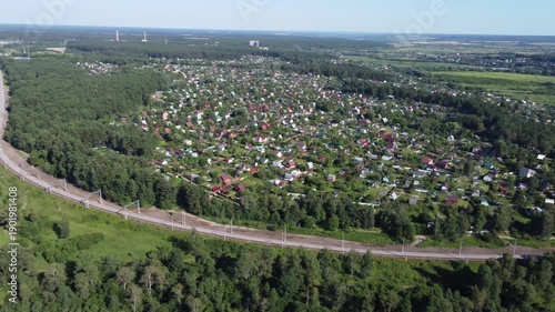 Aerial panorama of a vibrant dacha community and active railway snaking through verdant forests, with distant fields under a bright summer sky