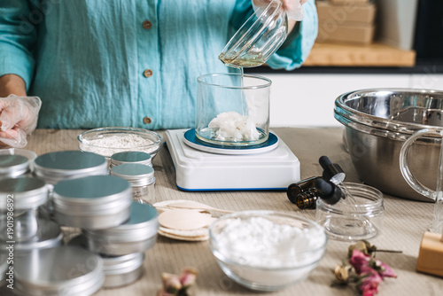 Fotografía Hand in glove pours liquid oil from bowl into beaker containing white solid butter on digital scale