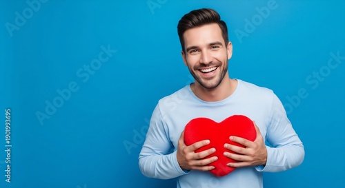 A smiling young man holds a red heart plush pillow against a vibrant blue studio background.