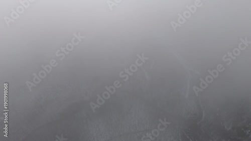 Aerial view of winter mountains with snow covered slopes as low clouds drift between ridges under sunny sky and heavy snow clouds.