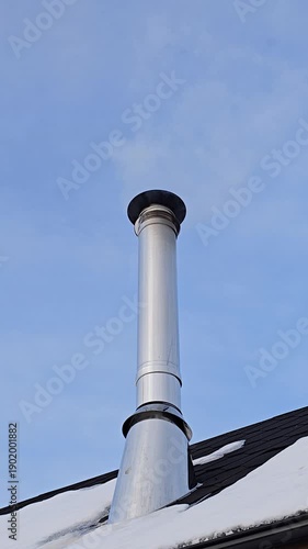 Close view of a silver chimney smoking on a small wooden house with snow on the roof clear blue sky and visible wood combustion fumes