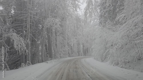 Vehicle moving on a narrow mountain road buried in fresh snow with white frozen trees around showing severe winter and dangerous road conditions.