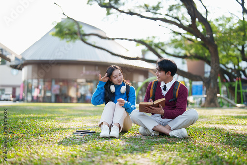Two young Asian students sitting outdoors on a green grass field, engaging in a lively discussion about their studies, sharing knowledge and developing new ideas together in an academic setting