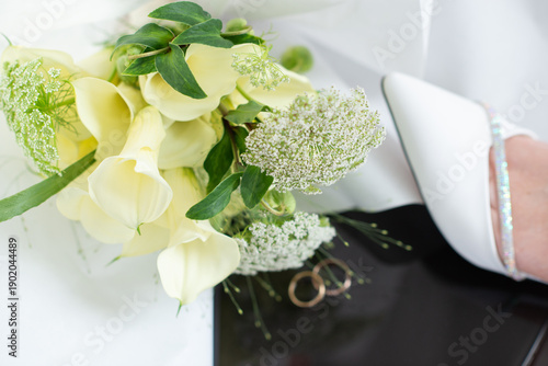 Bride resting her legs in elegant white heels near a window, with a wedding bouquet on a black table. Soft natural light, calm morning atmosphere, bridal preparation concept