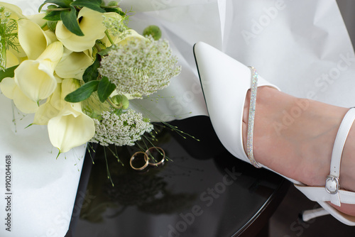 Bride resting her legs in elegant white heels near a window, with a wedding bouquet on a black table. Soft natural light, calm morning atmosphere, bridal preparation concept