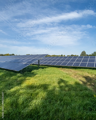 Solar panels stand in a field under a blue sky with clouds and sun during daytime hours in a rural area