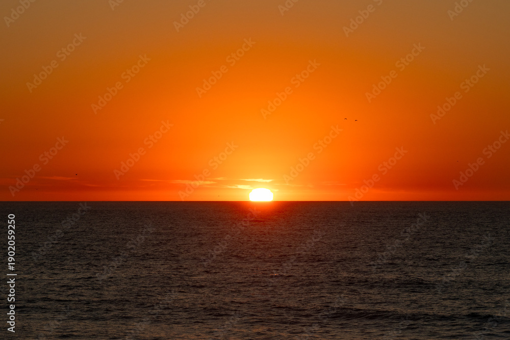 Fototapeta premium Fiery sun sinking below the ocean horizon during an intense orange sunset, Portugal, Ovar – Furadouro, 10.10.2025
