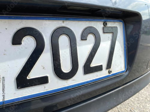 A detailed close-up of a front car license plate displaying the numbers 2027. The plate features a blue border and weathered dirt, suggesting travel, movement, and road life