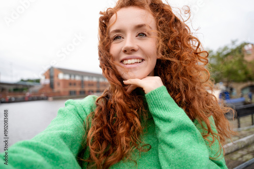 Obraz na płótnie Girl with curly red hair takes a selfie by the water on a cloudy day near a city