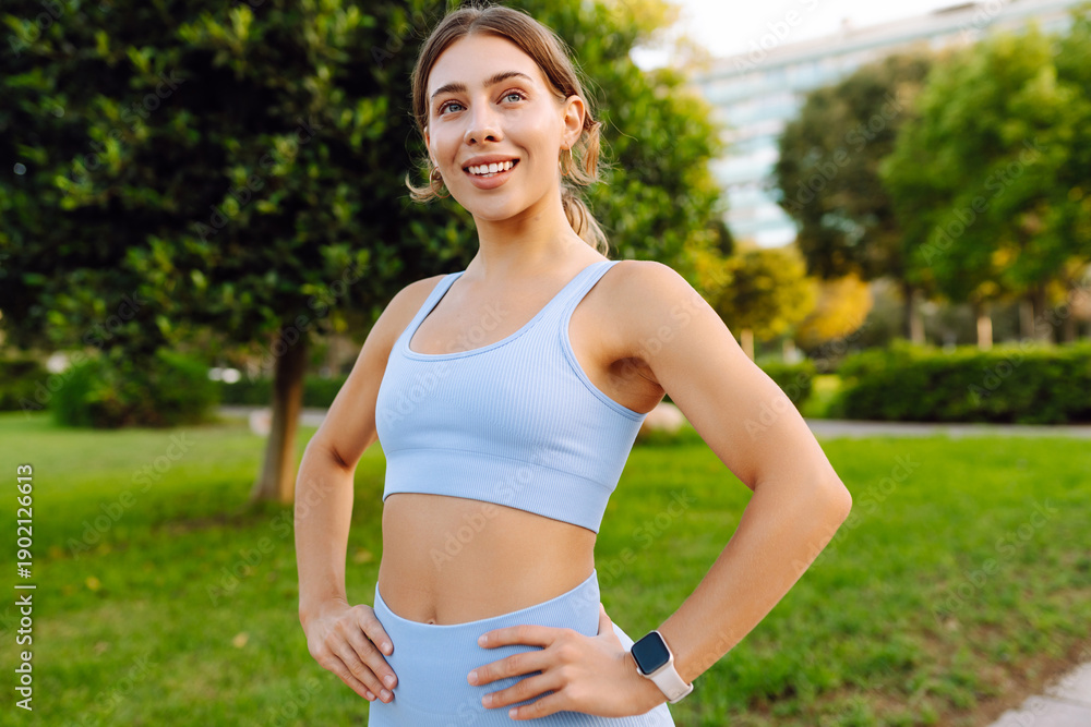Naklejka premium A smiling woman stands in a sunny park, stretching her arms. A beautiful woman in sportswear enjoys the sunny day before a workout. Sports concept, outdoors.