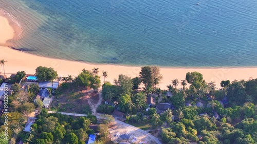 Top-down aerial view of Koh Phayam village coastline with sandy beach, wooden pier, fishing boats, tropical houses and clear turquoise sea.