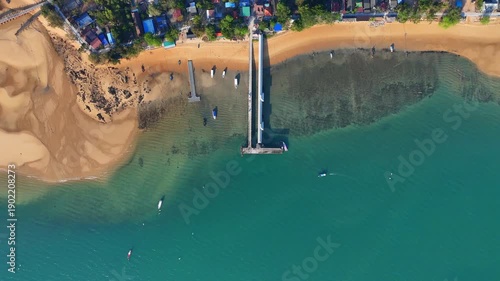 Top-down aerial view of Koh Phayam village coastline with sandy beach, wooden pier, fishing boats, tropical houses and clear turquoise sea.