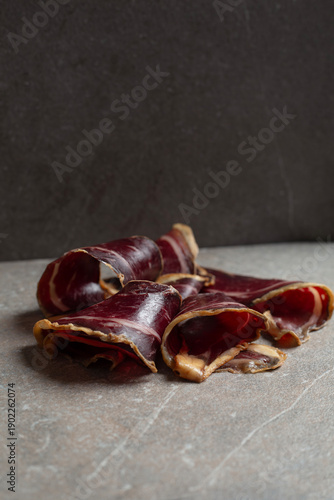 Elegant curls of Spanish cured beef cecina presented on a dark stone surface in a sophisticated low-light setting. Premium artisanal charcuterie with rich textures and deep tones, evoking luxury dinin