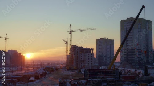 Stunning 4K timelapse of a sunrise over a bustling urban construction site, featuring cranes, high-rise buildings, and snowy terrain under a golden sky.