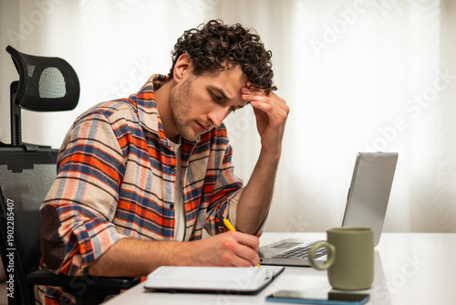Wallpaper Mural Serious young businessman thinking, taking notes and using laptop while working remotely at his cozy home. Torontodigital.ca
