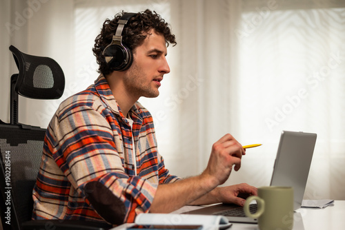 Wallpaper Mural Modern young man using wireless headphones and laptop while participating in e-learning session from the comfort of his home.	 Torontodigital.ca