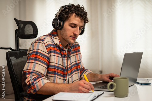 Wallpaper Mural Focused young man wearing wireless headphones, working on his laptop at home while taking notes on a notebook.	 Torontodigital.ca