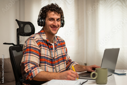 Wallpaper Mural Portrait of happy young man wearing wireless headphones, working on his laptop at home while taking notes on a notebook. Torontodigital.ca