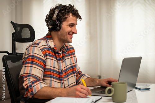 Wallpaper Mural Happy young man wearing wireless headphones, working on his laptop at home while taking notes on a notebook. Torontodigital.ca