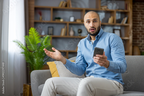 Fotografi Surprised man sitting on sofa, holding mobile phone, feeling disbelief and confu