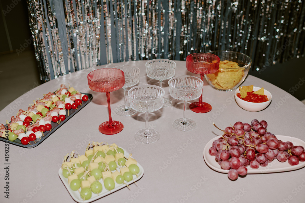 Fototapeta premium Table displaying empty crystal glasses, red and clear goblets, plates with green and red grapes, skewered fruit appetizers, bowl of potato chips, bowl of red jelly candies