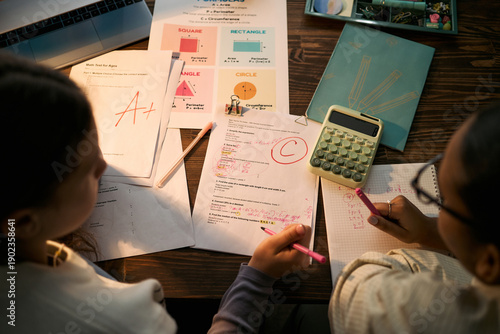 Two teenage girls, one Black and one Caucasian, studying together at desk, reviewing graded math assignments with calculator and worksheets, discussing solutions during tutoring lesson