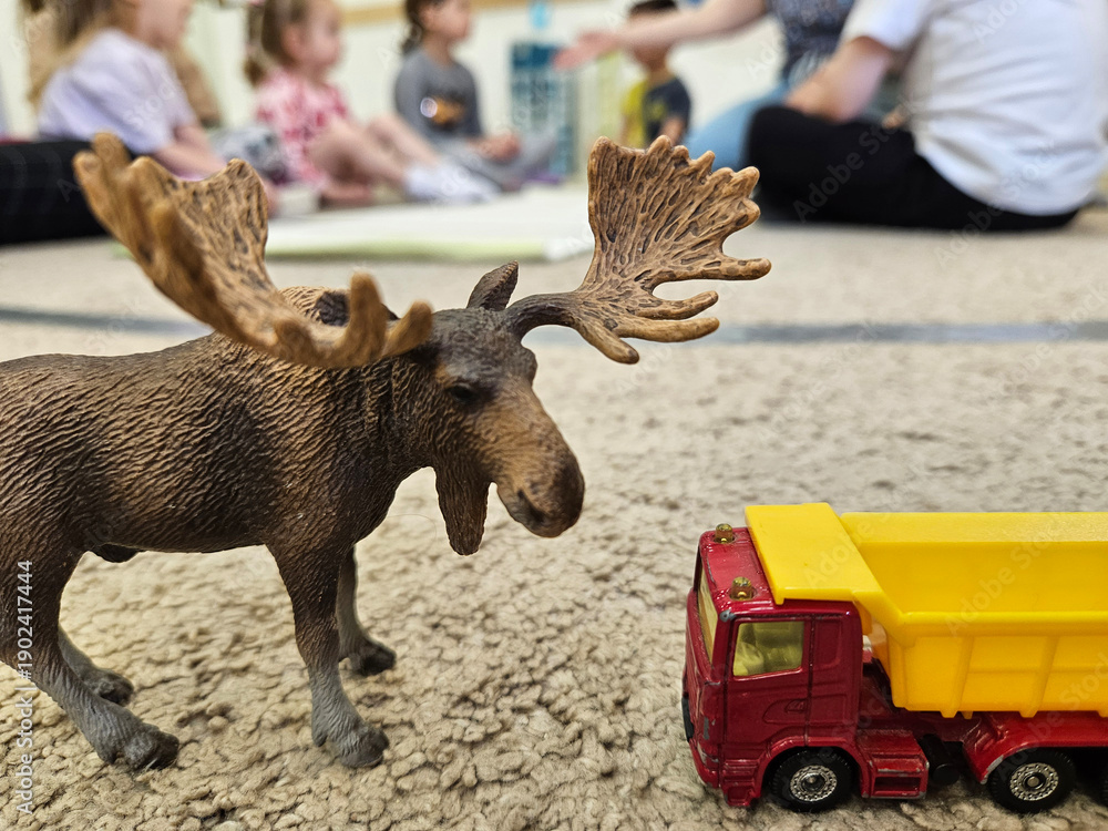 Fototapeta premium Toy truck is next to a toy moose. The truck is red and yellow. The moose is brown and has a large head. The scene takes place on a carpeted floor, with the children sitting in kindergarten
