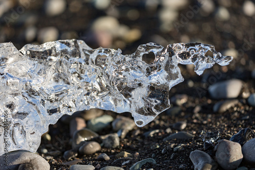 A sculpted fragment of clear ice rests on the dark pebbled shore of Diamond Beach near Jökulsárlón glacier lake as gentle afternoon light glows across Southern Iceland’s volcanic coastline