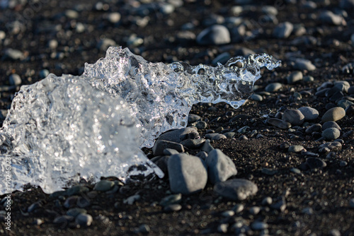 A jagged crystal of ice glows on the dark pebbled shore of Diamond Beach near Jökulsárlón glacier lake as soft evening light brushes Southern Iceland’s rugged volcanic coastline