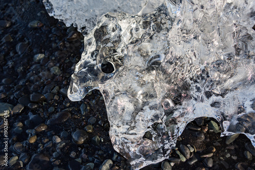 A sculpted shard of clear ice rests on the dark pebbled shore of Jökulsárlón glacier lake as soft morning light reveals its delicate patterns against Southern Iceland’s rugged ground