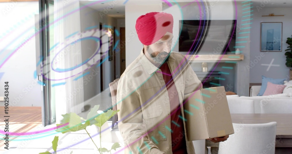 Fototapeta premium Mid-adult Indian man wearing red turban and beige jacket holding cardboard box at home, with plant