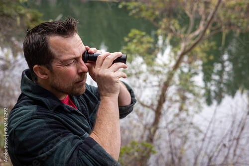 A close-up side profile of a middle-aged man with a beard using a handheld black monocular to observe nature in a lush forest setting with a lake in the background.