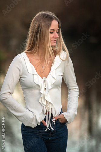 Young woman with long blonde hair wearing a white blouse and blue jeans stands by a reflective body of water in a natural outdoor setting
