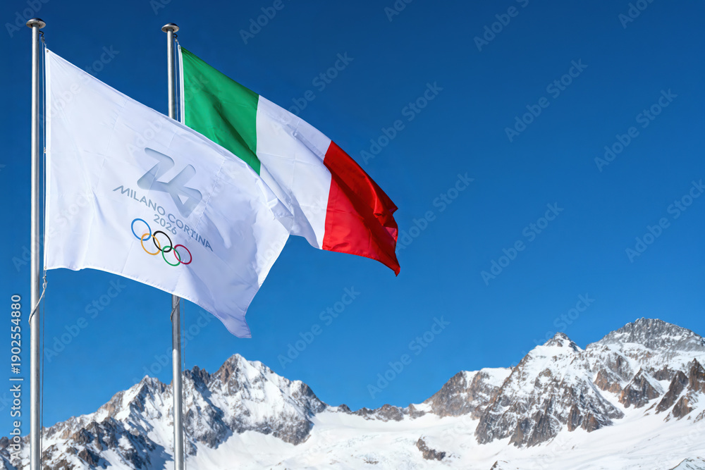 Fototapeta premium MILAN, ITALY, DECEMBER 12, 2025. The Italian national flag and the Milano Cortina 2026 Winter Olympics flag with the iconic rings wave over the snowy peaks of the Italian Alps in the sun Ai generated