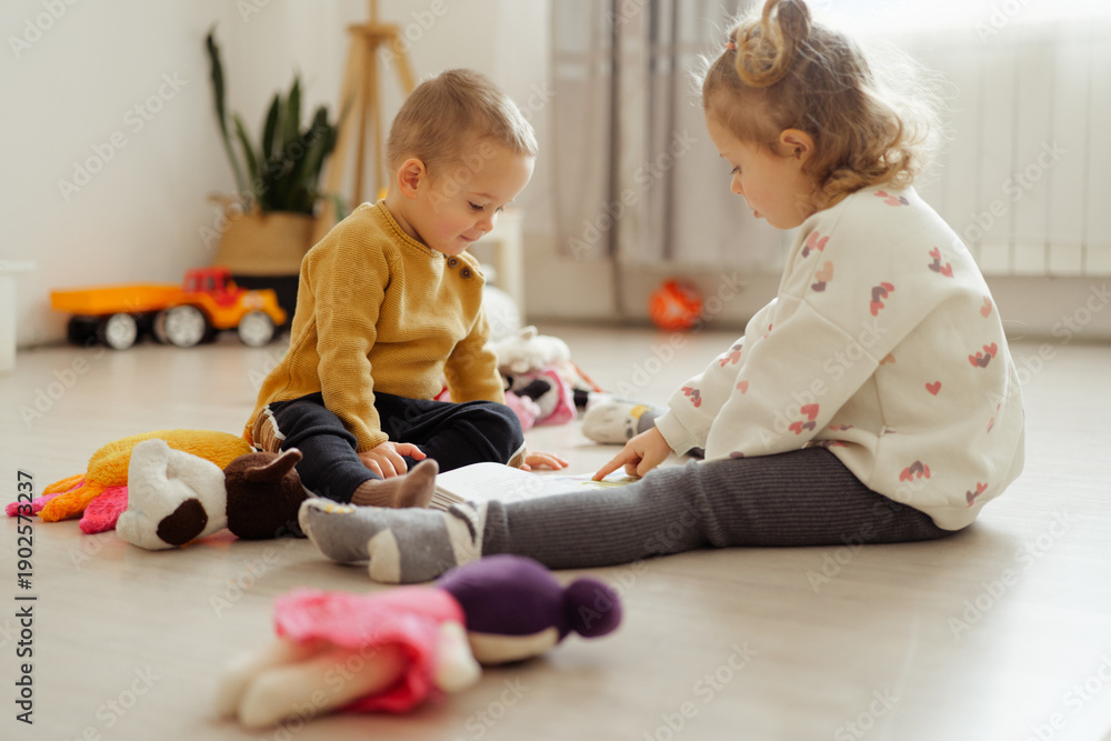 Naklejka premium Two young children sit on the floor with toys around them. They focus on a book laid out in front of them in a well-lit room during the daytime