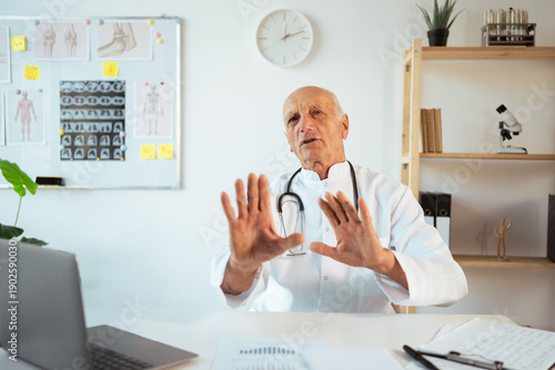 senior male doctor wearing white coat work on laptop in hospital clinic, say no thank you with hands on camera